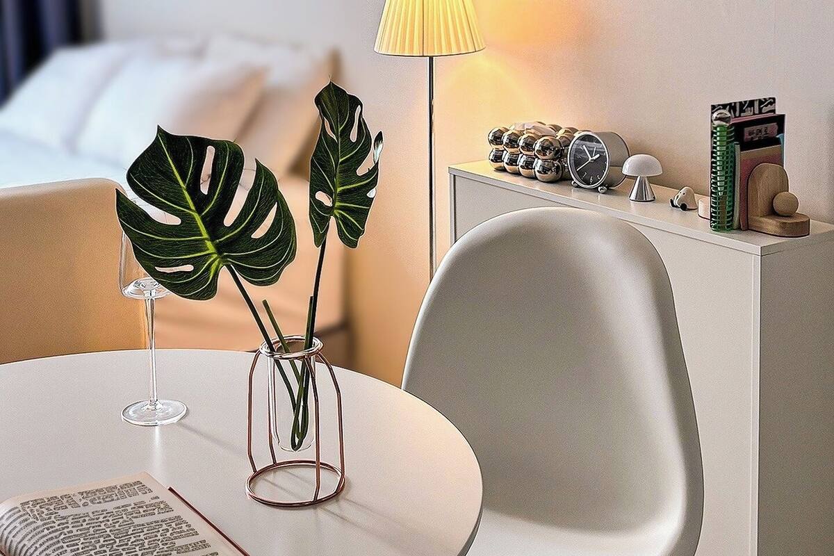 A modern hotel room with a round dining table, a bedside lamp, a plant, and a bedside clock.