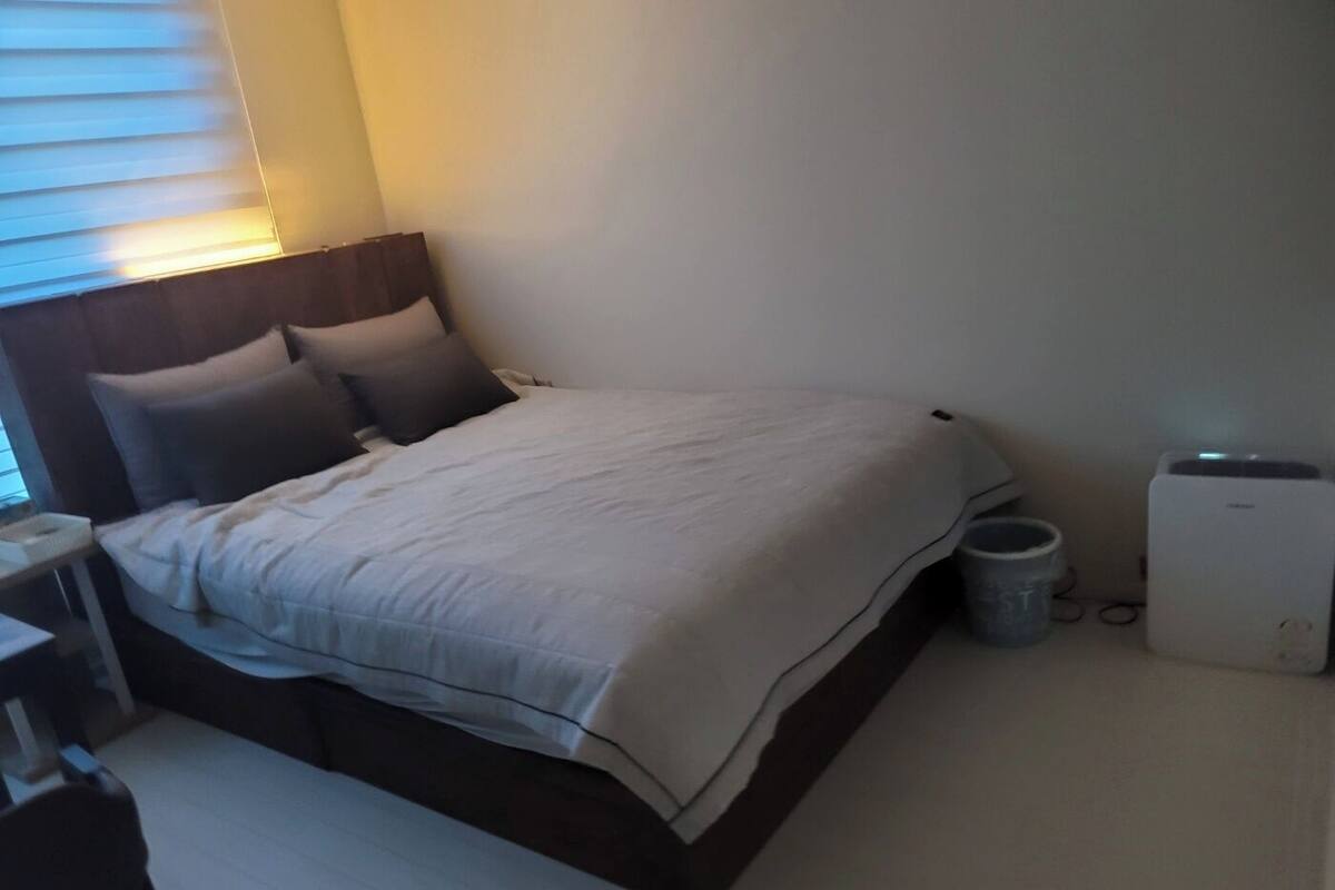 A neatly made bed with a white comforter and pillows, a bedside table with a lamp, and a small trash bin.