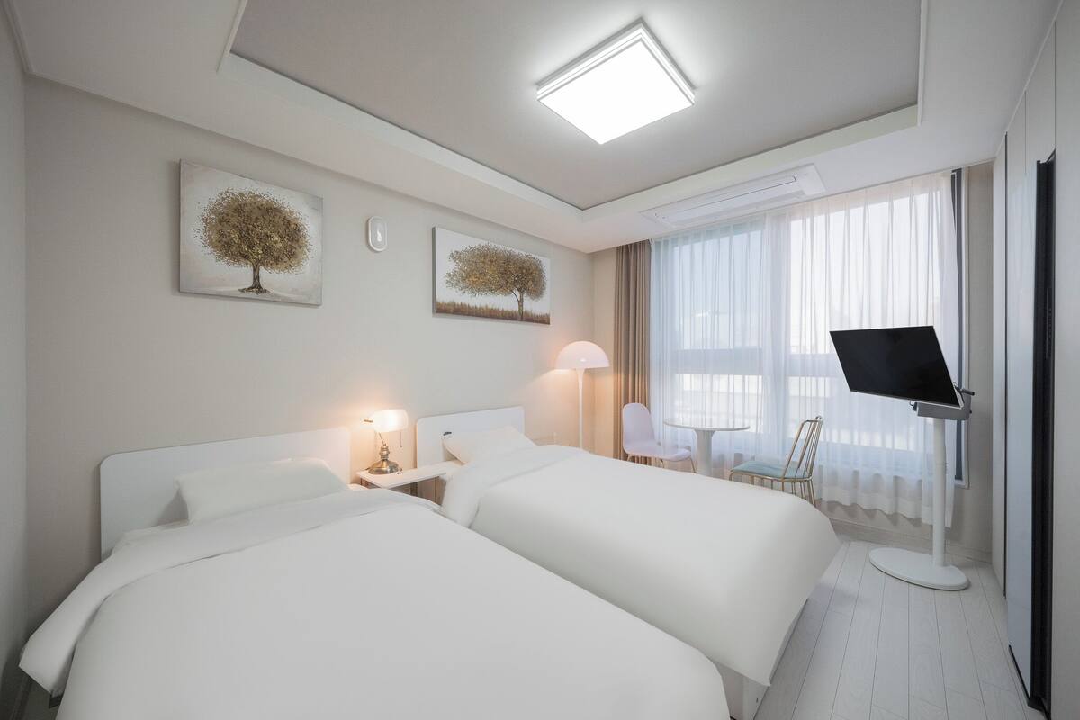 A hotel room with two beds, a desk with a chair, a TV on a stand, and artwork on the wall.