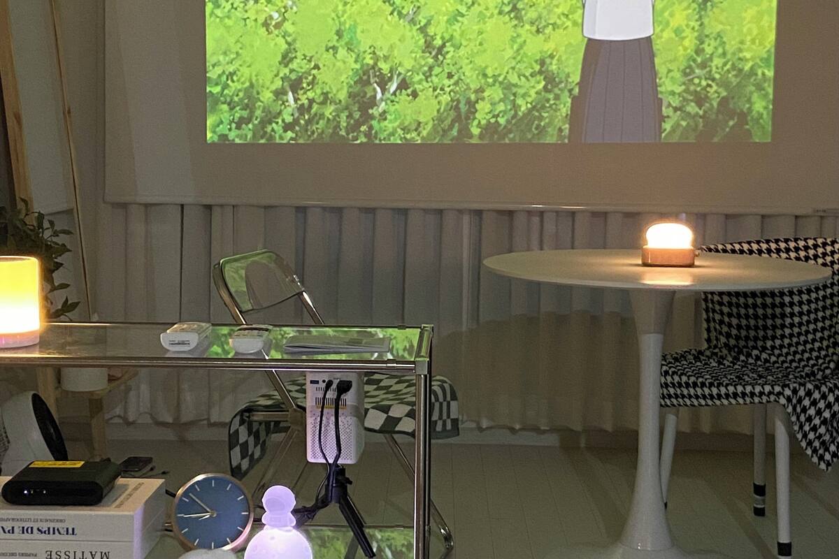 A room with a projection screen displaying an animated character, a table with a lamp, a chair, and a clock.
