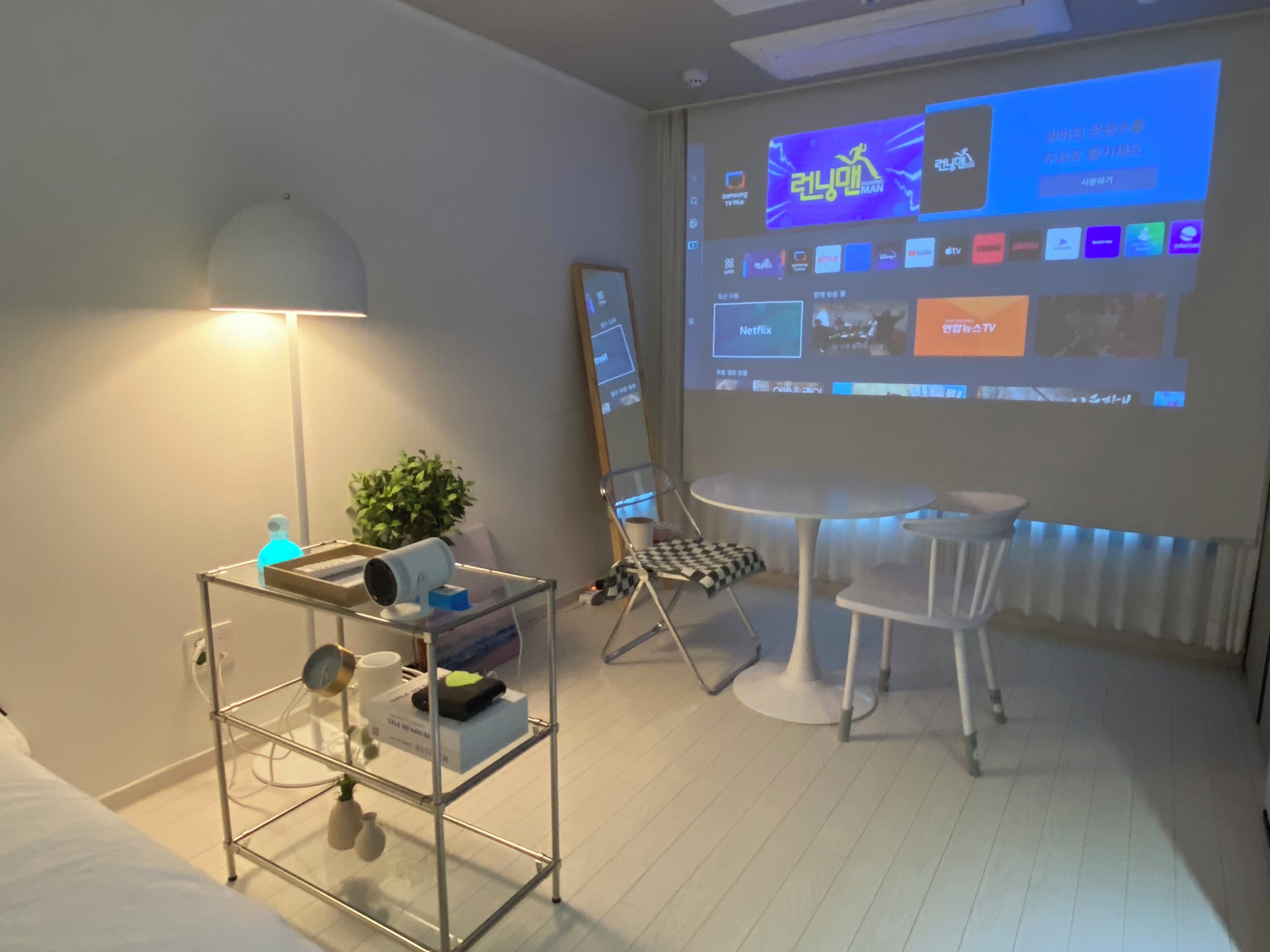 Ein modernes Hotelzimmer mit Bett, Schreibtisch mit Lampe, kleinem Tisch, Stuhl und einem Projektorbildschirm, der eine Fernsehsendung zeigt.