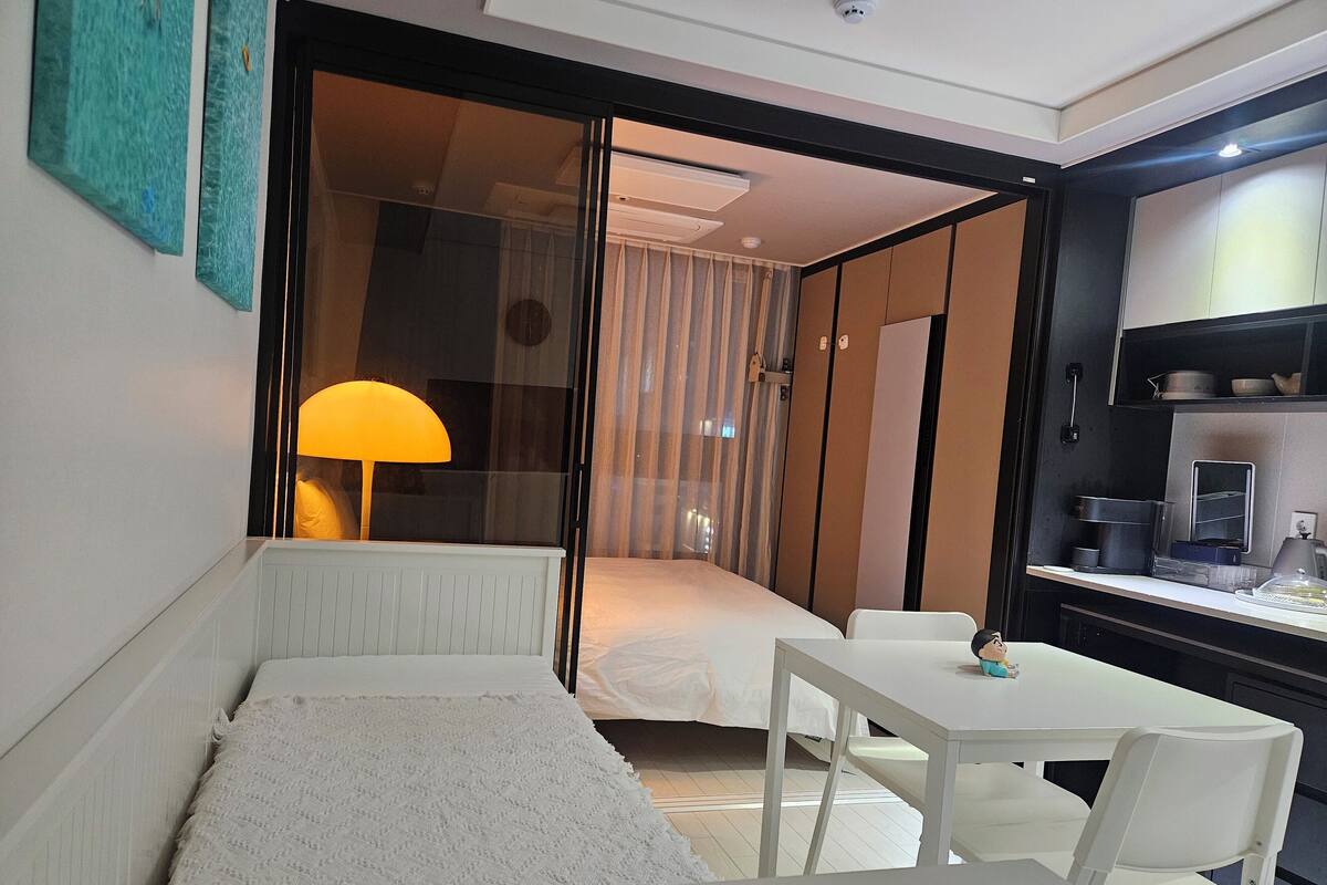 A compact hotel room with a bed, a small sitting area with a table and chairs, a desk with a lamp, and a kitchenette.