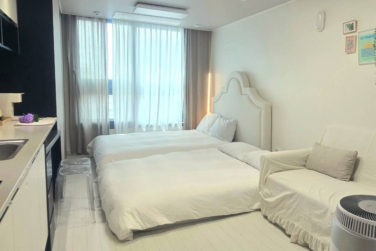 A hotel room with a double bed, a bedside table, a chair, a small desk with a sink, and a window with curtains.