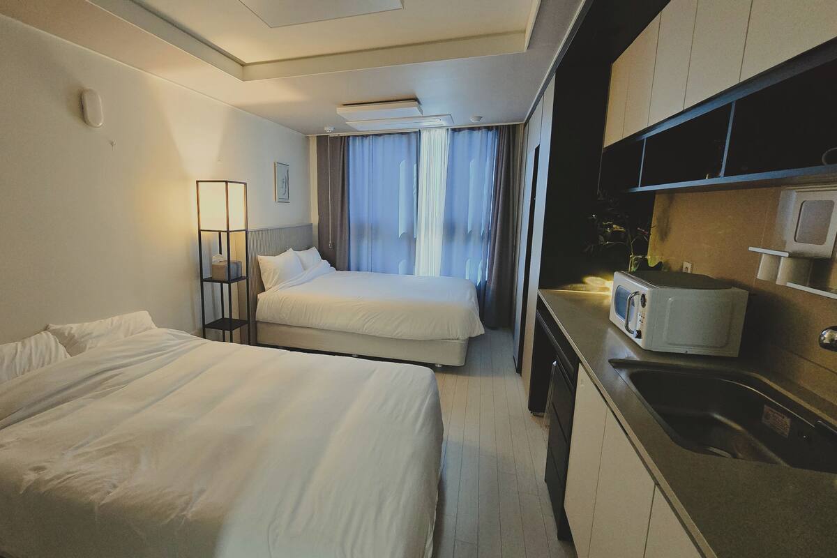 A hotel room with two beds, a kitchenette, and a window with curtains.