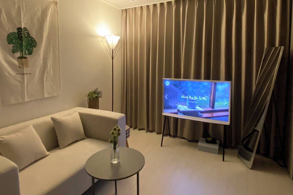 A modern living room with a beige sofa, a small round table with a vase, a floor lamp, and a flat-screen TV on a stand.