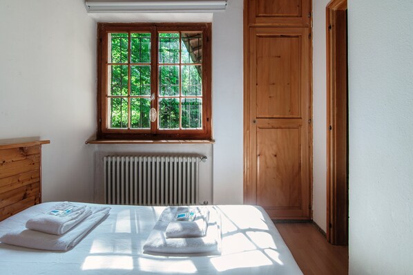 A bedroom with a wooden headboard, a window with a view of greenery, a radiator, and a neatly made bed with towels.