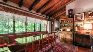 A wooden dining area with a table and chairs, a red rug, and a view of greenery through large windows. - Casa al Fiume (Sonogno)