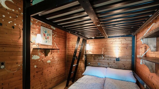 A wooden cabin room with a bunk bed, a ladder, and a wall-mounted shelf with a lamp.