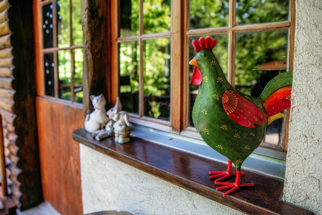 A rooster figurine on a window sill with a view of greenery outside.
