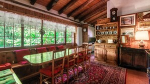 Un comedor rústico de madera con una mesa y sillas, un banco con cojines, un tapete estampado y una zona de cocina con armarios de madera y un reloj.