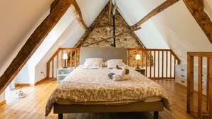 Un dormitorio acogedor en el ático, con techo inclinado, vigas de madera, una cama grande con ropa de cama estampada, lámparas de noche y un pequeño aparador blanco.