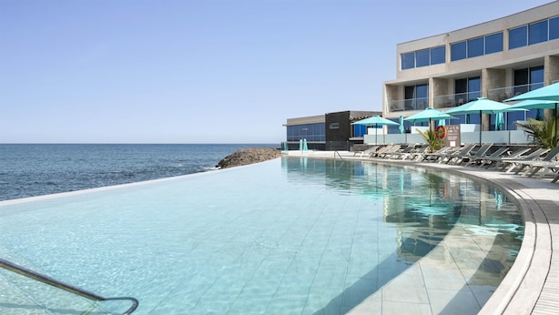Ein klares Schwimmbecken mit Liegestühlen und Sonnenschirmen, ein Gebäude mit großen Fenstern und Blick aufs Meer.