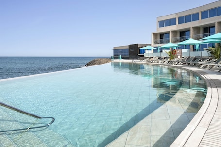 A clear swimming pool with lounge chairs and umbrellas, a building with large windows, and a view of the sea.