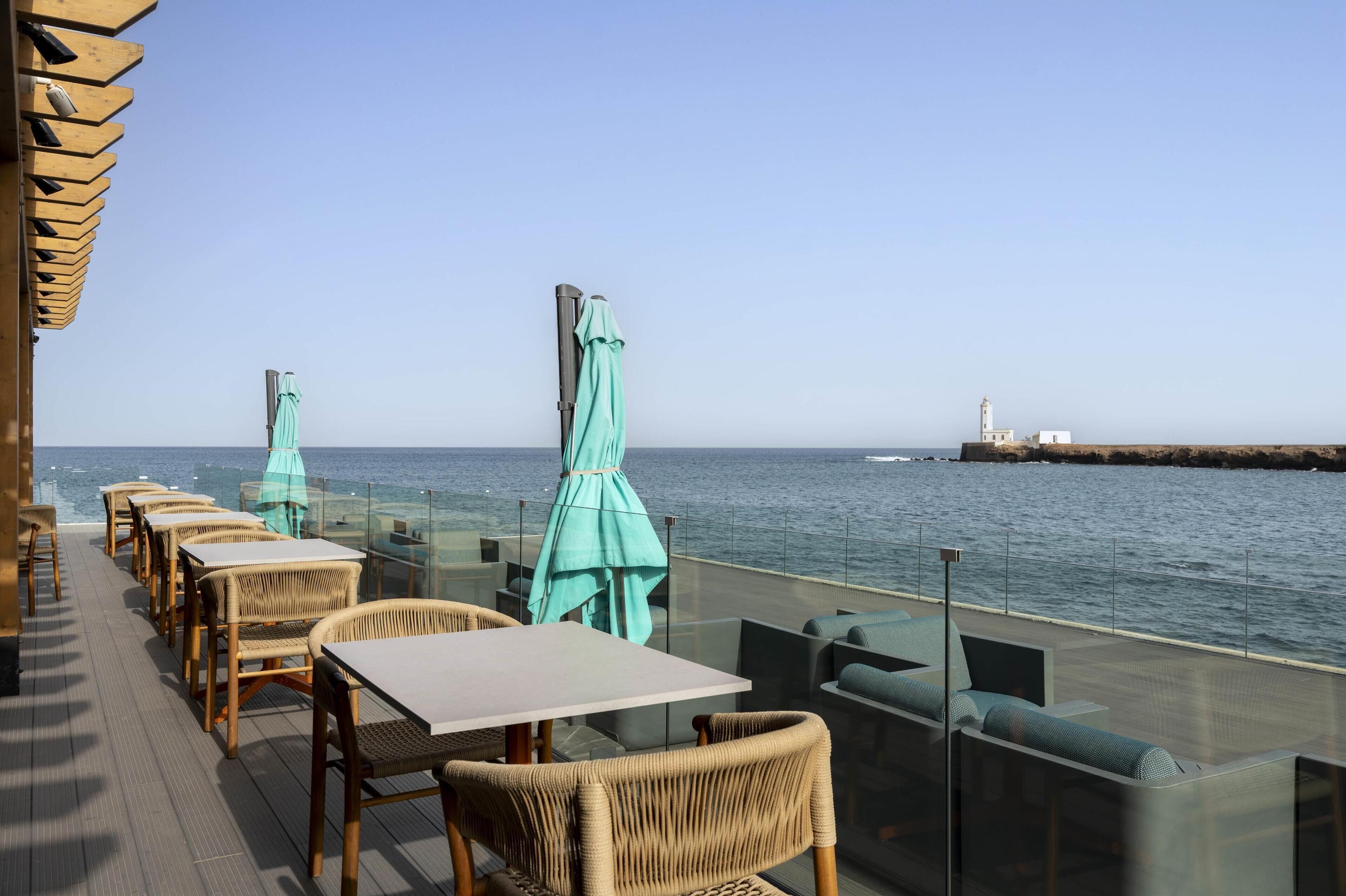 A seaside restaurant with wicker tables and chairs, turquoise umbrellas, and a view of the ocean and a lighthouse.