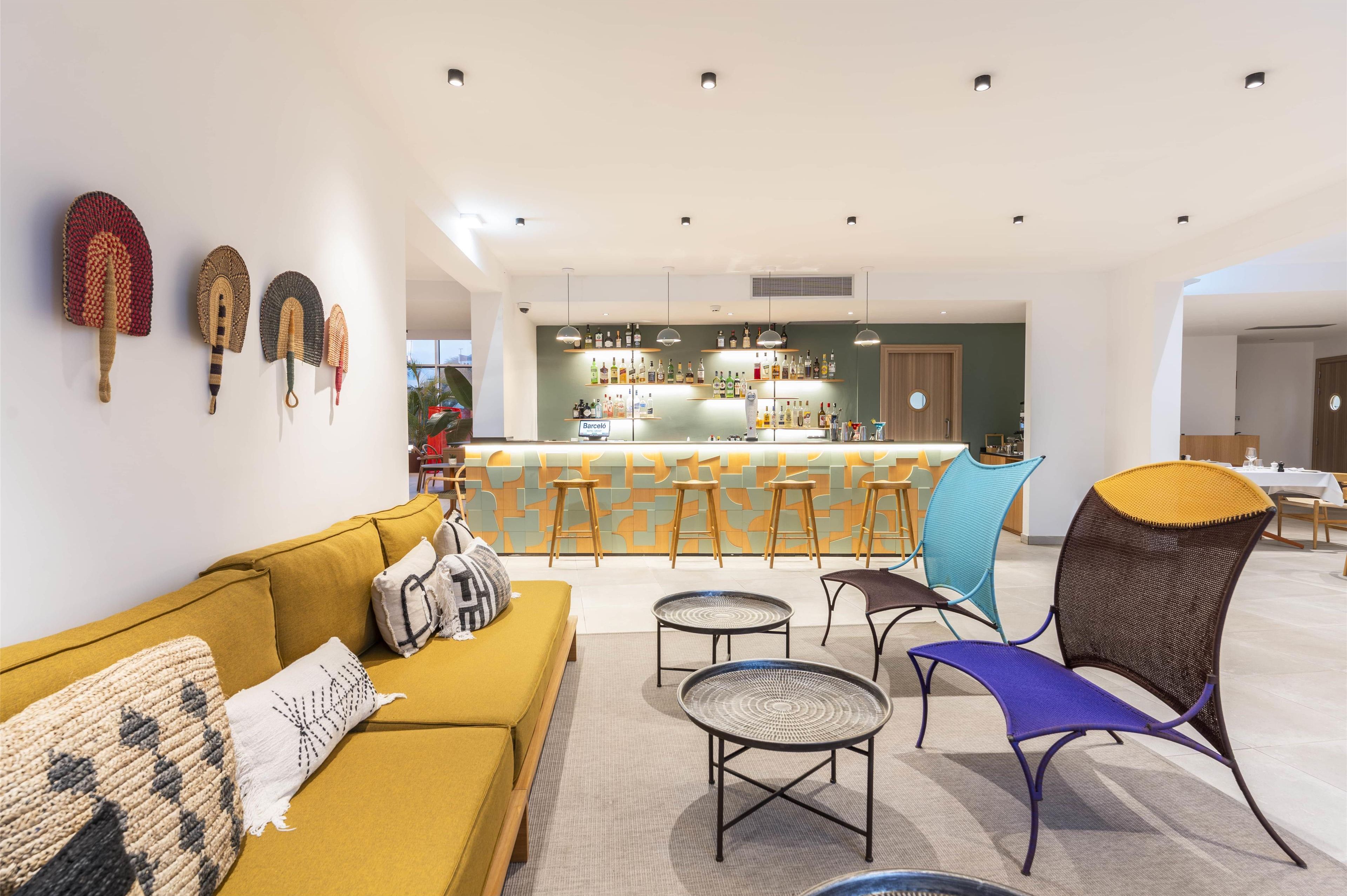 A modern living room with a yellow sofa, round tables, and a bar area with stools and shelves of bottles.