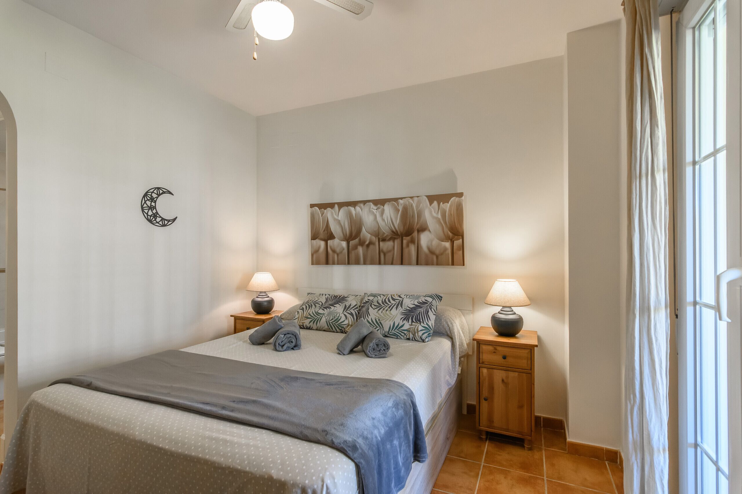 A bedroom with a bed, two bedside tables with lamps, a wall clock, and a window with curtains.