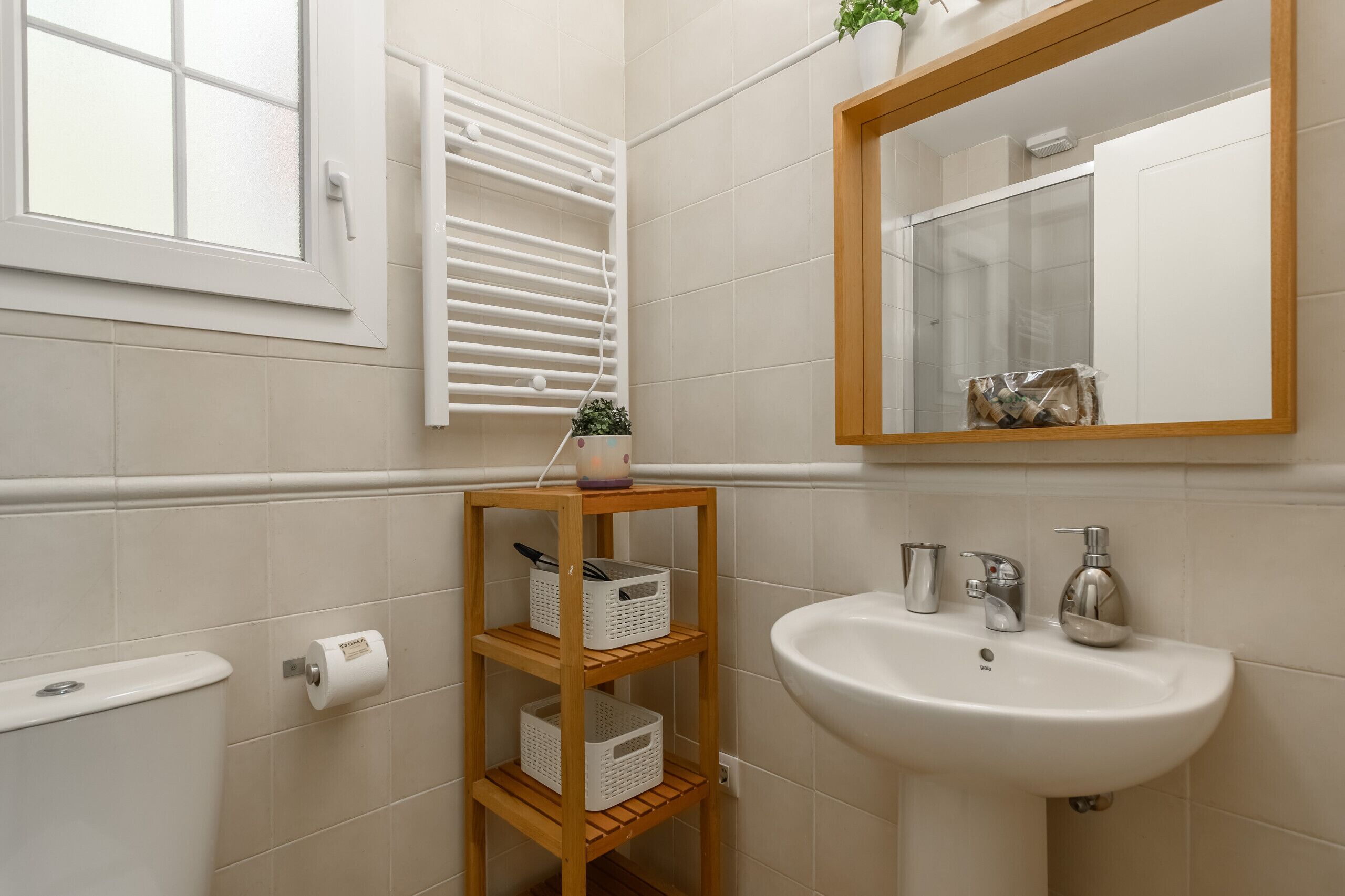Une salle de bain avec un lavabo blanc, un miroir, une fenêtre avec des stores et une étagère avec des paniers.