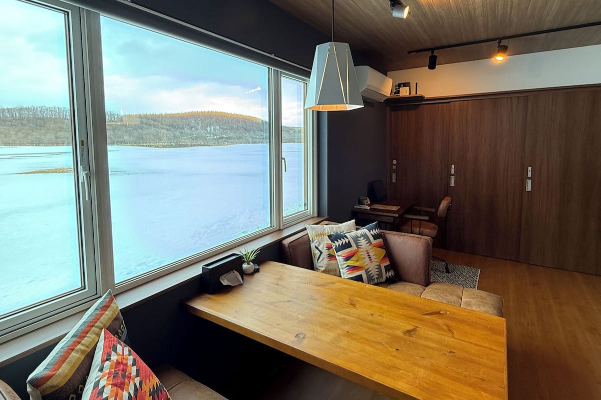 A modern interior with a large window overlooking a lake, a wooden bench, and a dining area with a table and chairs.