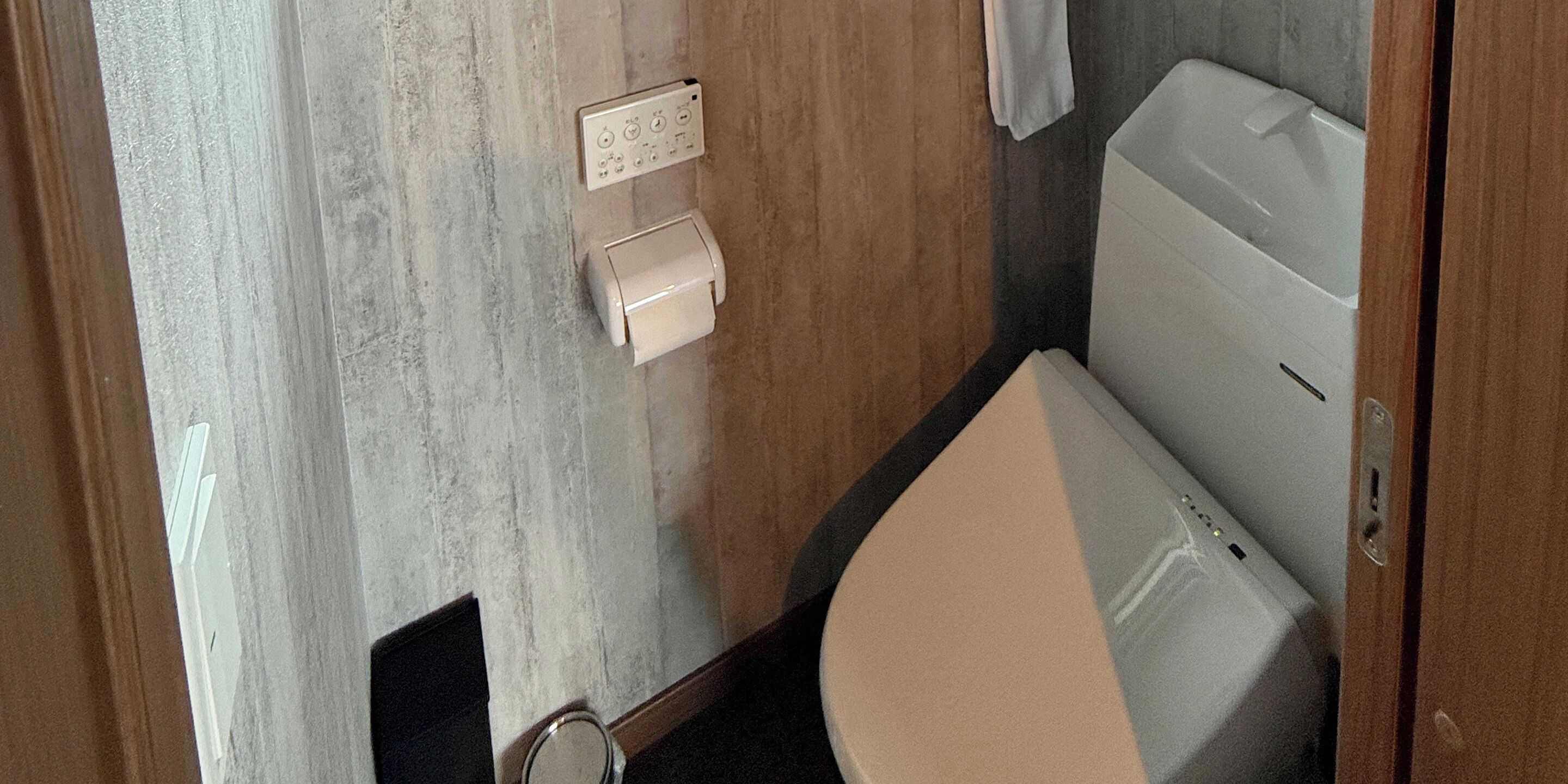 A compact bathroom with a toilet, a wall-mounted flush handle, and a small trash bin.