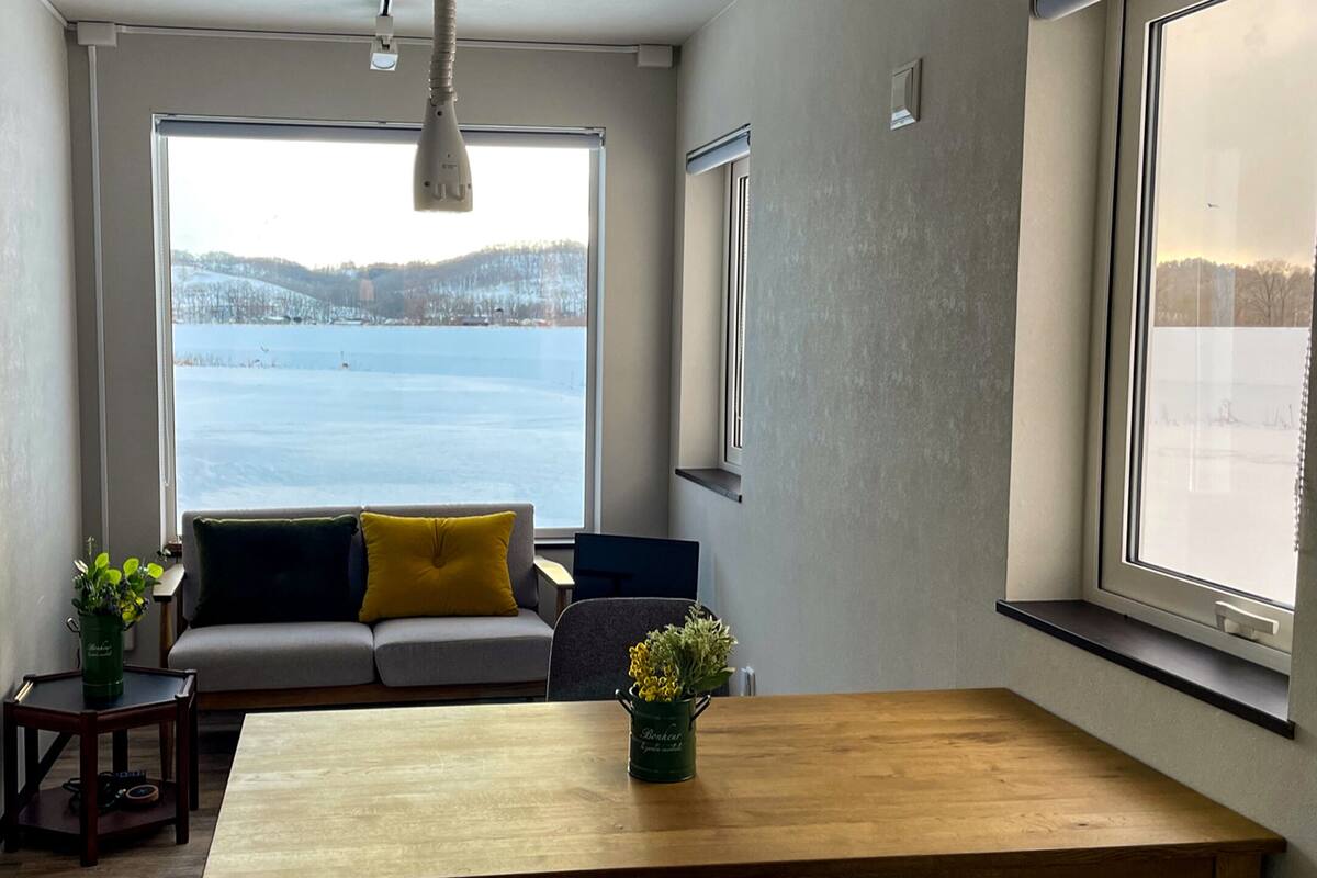 A room with a large window overlooking a snowy landscape, a sofa with cushions, a wooden table, and a small plant on the table.