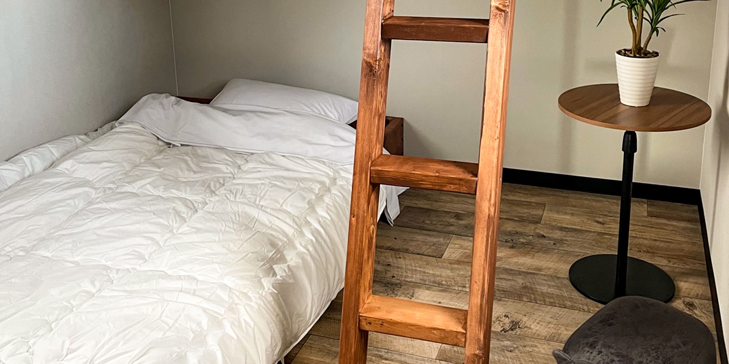 Une chambre avec une échelle en bois appuyée contre un lit, une table ronde en bois sur laquelle est posée une plante en pot, et une chaise grise.