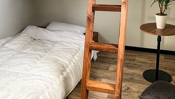 Une chambre avec une échelle en bois appuyée contre un lit, une table ronde en bois sur laquelle est posée une plante en pot, et une chaise grise.