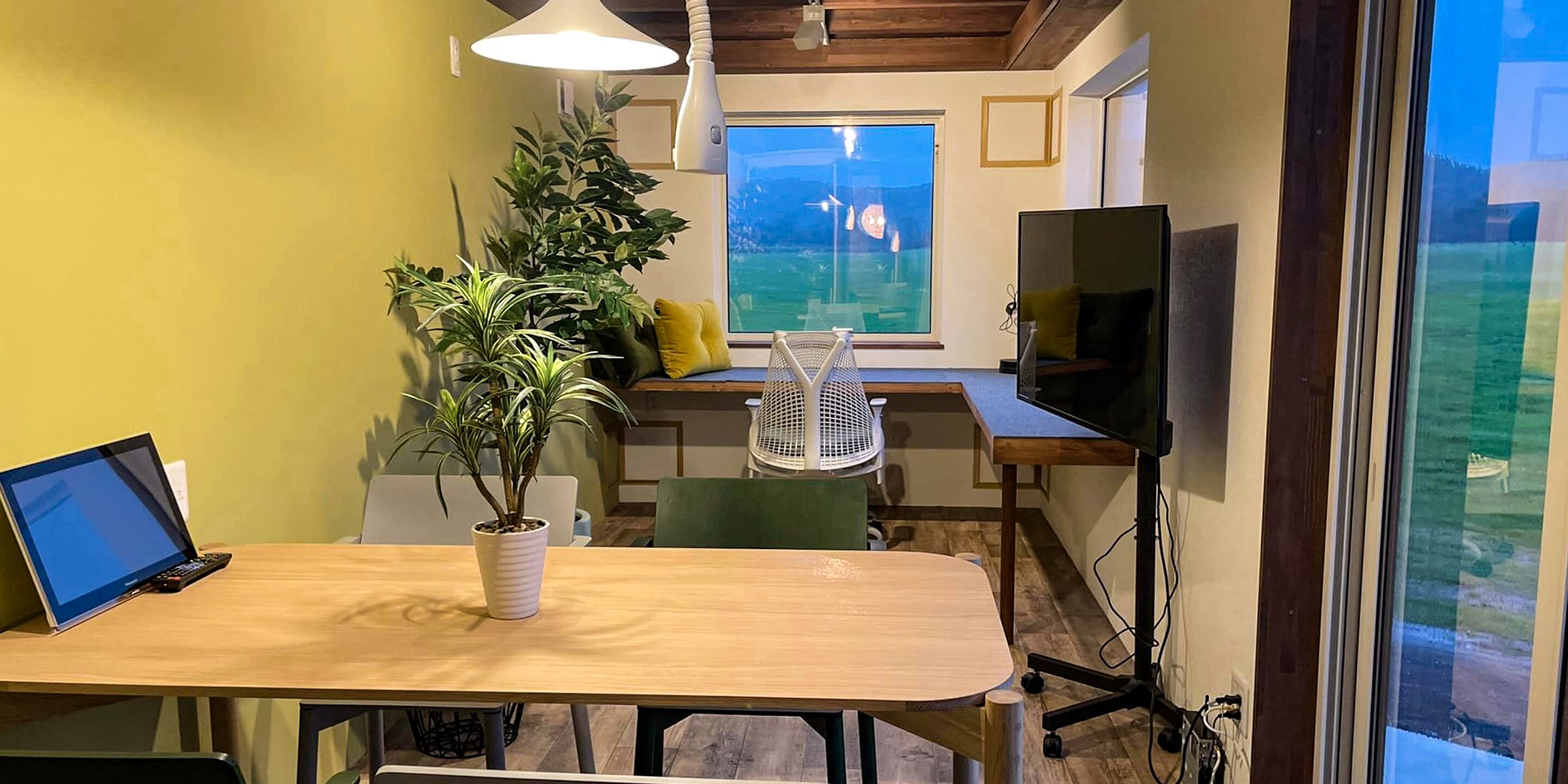 Een moderne kamer met een houten tafel, een laptop, een potplant, een groot raam en een televisie.
