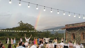Un événement en plein air, illuminé de guirlandes lumineuses, où les invités sont assis à des tables, avec un arc-en-ciel en arrière-plan.