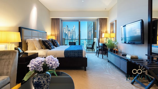 A modern hotel room with a large bed, a desk, a chair, a TV, and a view of a cityscape. - Luxe High Floor In Address Marina.36 (Dubai)