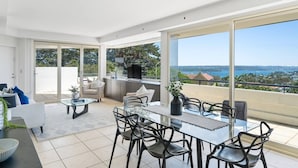 Una sala de estar moderna con una mesa y sillas de vidrio, un sofá y un televisor. Cuenta con un balcón con vista al mar.