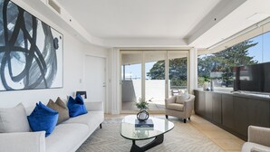 Una sala de estar moderna con una mesa de centro de vidrio, un sofá con cojines azules, un sillón color beige y un cuadro abstracto grande en la pared.