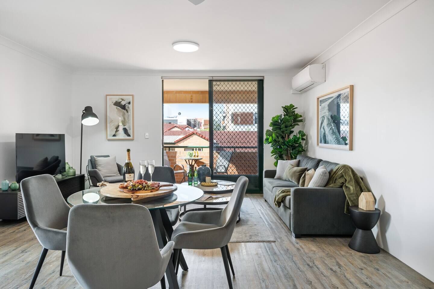 A modern living room with a dining area, grey furniture, a TV, and a balcony with a view of buildings.