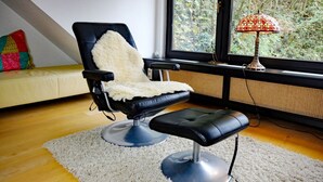 Une pièce moderne avec un fauteuil en cuir noir, un petit tabouret, un tapis à motifs et une vue sur les arbres à travers une grande fenêtre.