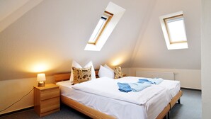 A bedroom with a sloped ceiling, a bed with white bedding, two bedside lamps, and two windows with wooden frames.
