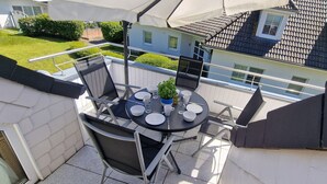A balcony with a table set for four, surrounded by metal chairs, overlooking a residential area with a house and well-maintained lawn.