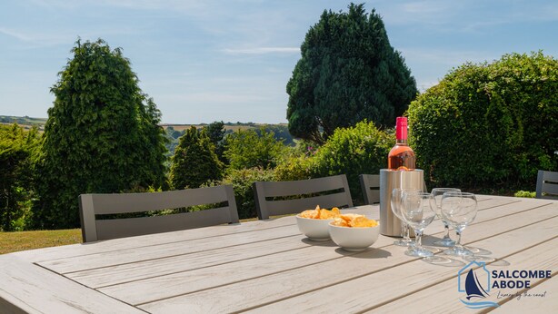 A wooden table with a bottle of wine, glasses, and bowls of chips in an outdoor setting with trees and hills in the background. - Detached 4 bed Coastal Haven  | Garden  |  Parking (Salcombe)