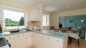 A kitchen with a large window, a dining area with a table set for two, and a view of the outdoors. - Detached 4 bed Coastal Haven  | Garden  |  Parking (Salcombe)