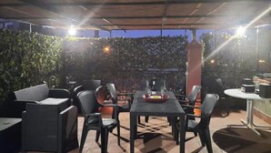 An outdoor seating area with tables and chairs, a covered section, and a backdrop of greenery. - Casa Leto 5 (Letojanni)