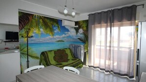 A room with a dining table, chairs, a sofa with a floral cushion, a TV on a shelf, and a mural of a beach scene. - Casa Leto 5 (Letojanni)