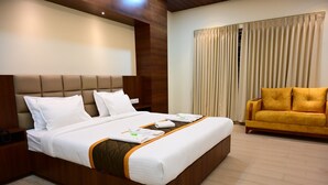 A hotel room with a large bed, a wooden headboard, a wooden nightstand, a yellow sofa, and a window with curtains. - Unnati Stays Inn, VIJAYAPUR (VIJAYAPUR)