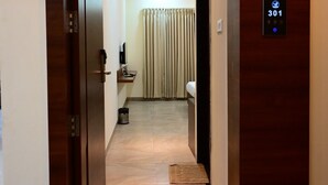A hotel hallway with a wooden door, a digital elevator control panel, and a view into a room with a bed and curtains. - Unnati Stays Inn, VIJAYAPUR (VIJAYAPUR)