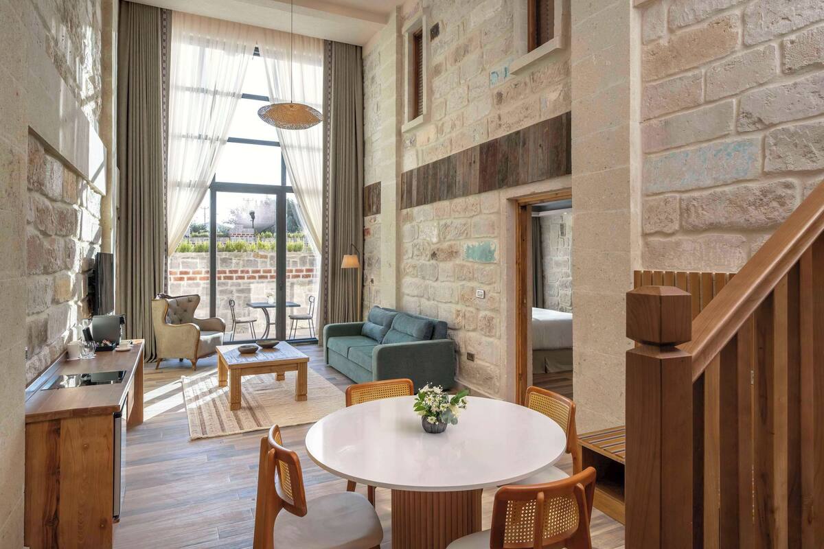 Ein gemütlicher Wohnbereich mit rundem Tisch, Stühlen und einer Couch. Es gibt eine Küchenzeile mit Spüle und einem Fenster mit Blick auf einen Garten.