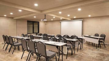 Un salón de conferencias con varias mesas y sillas, equipado con proyectores y pantallas, y suelo de baldosas.