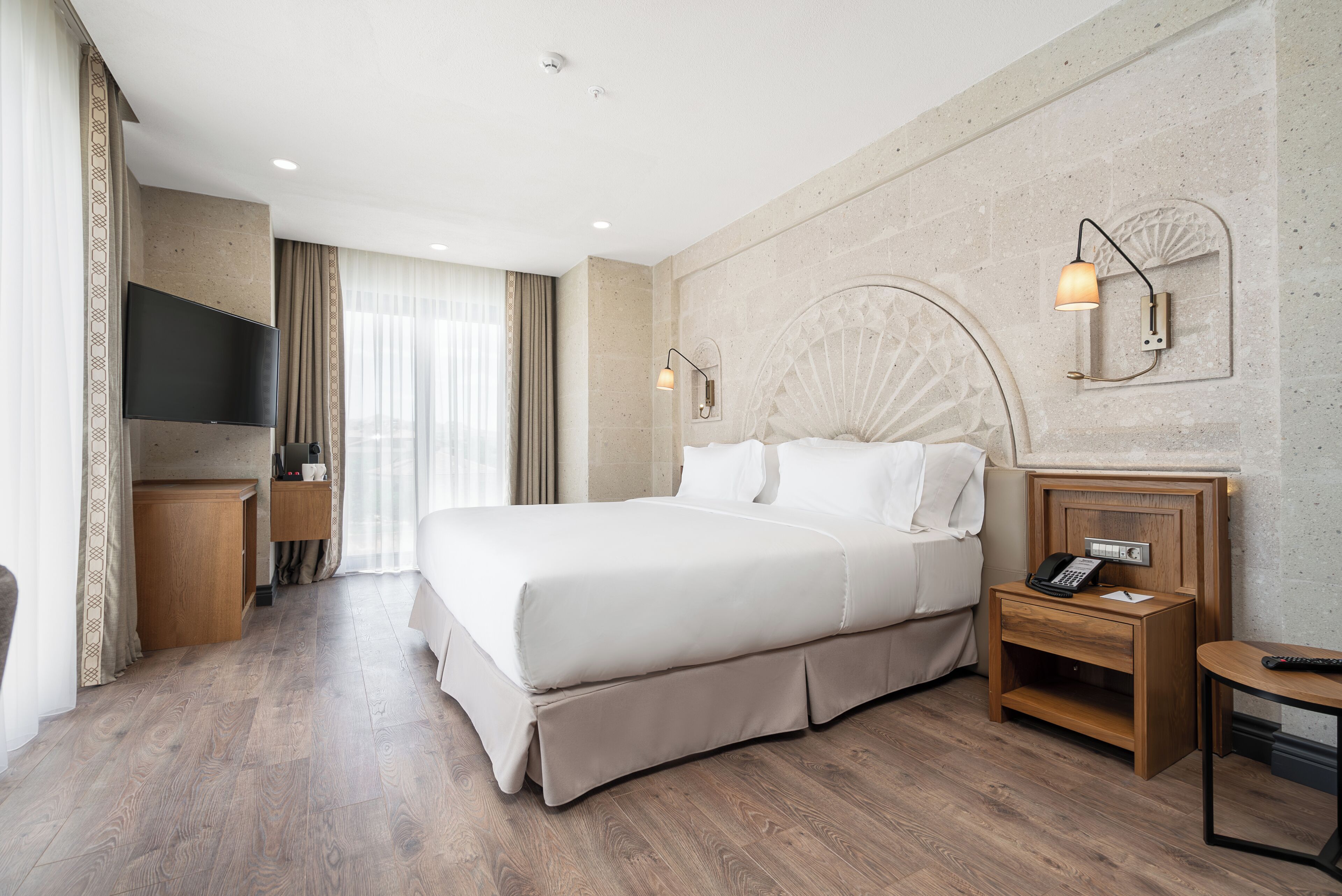 Una habitación de hotel con una cama grande, televisión, mesita de noche de madera y una lámpara de pared.
