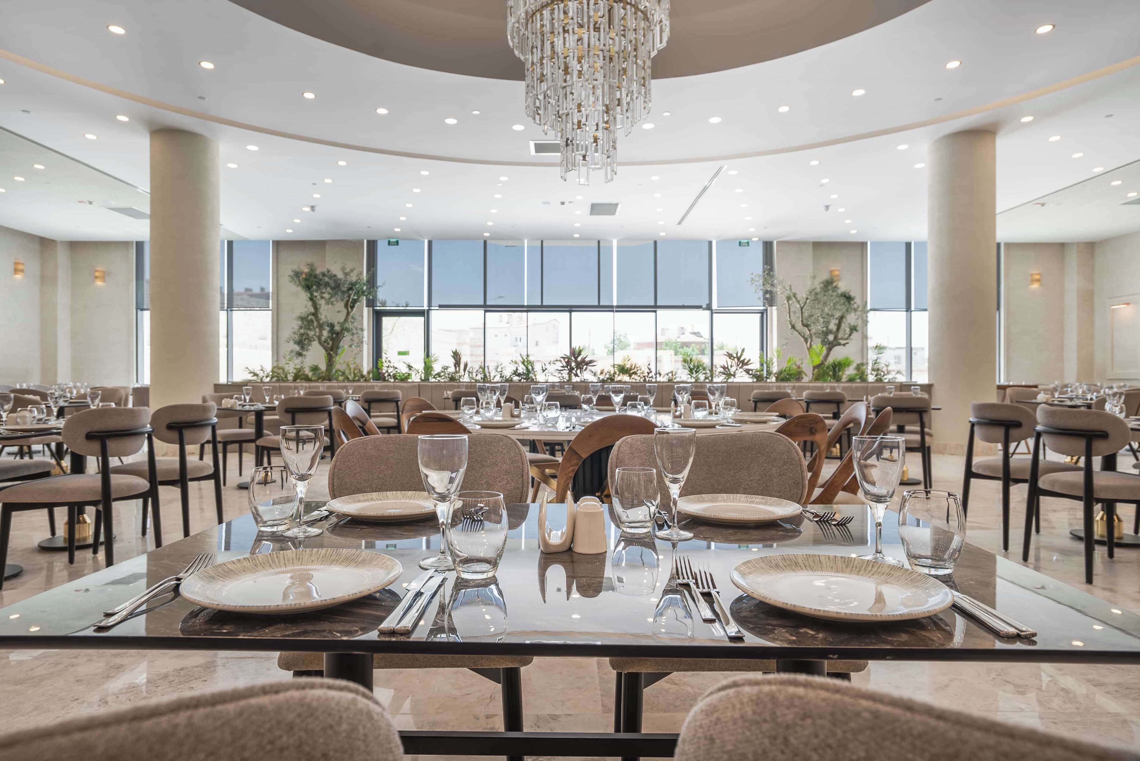 Uma sala de jantar ampla com várias mesas preparadas para o serviço, grandes janelas e um candelabro.