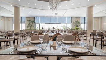Uma sala de jantar ampla com várias mesas preparadas para o serviço, grandes janelas e um candelabro.