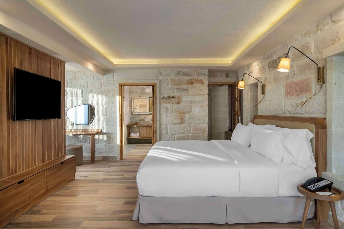Ein Schlafzimmer mit einem großen Bett, Holzvertäfelung an einer Wand, einem Flachbildfernseher, einem Nachttisch mit Telefon und Steinwänden.