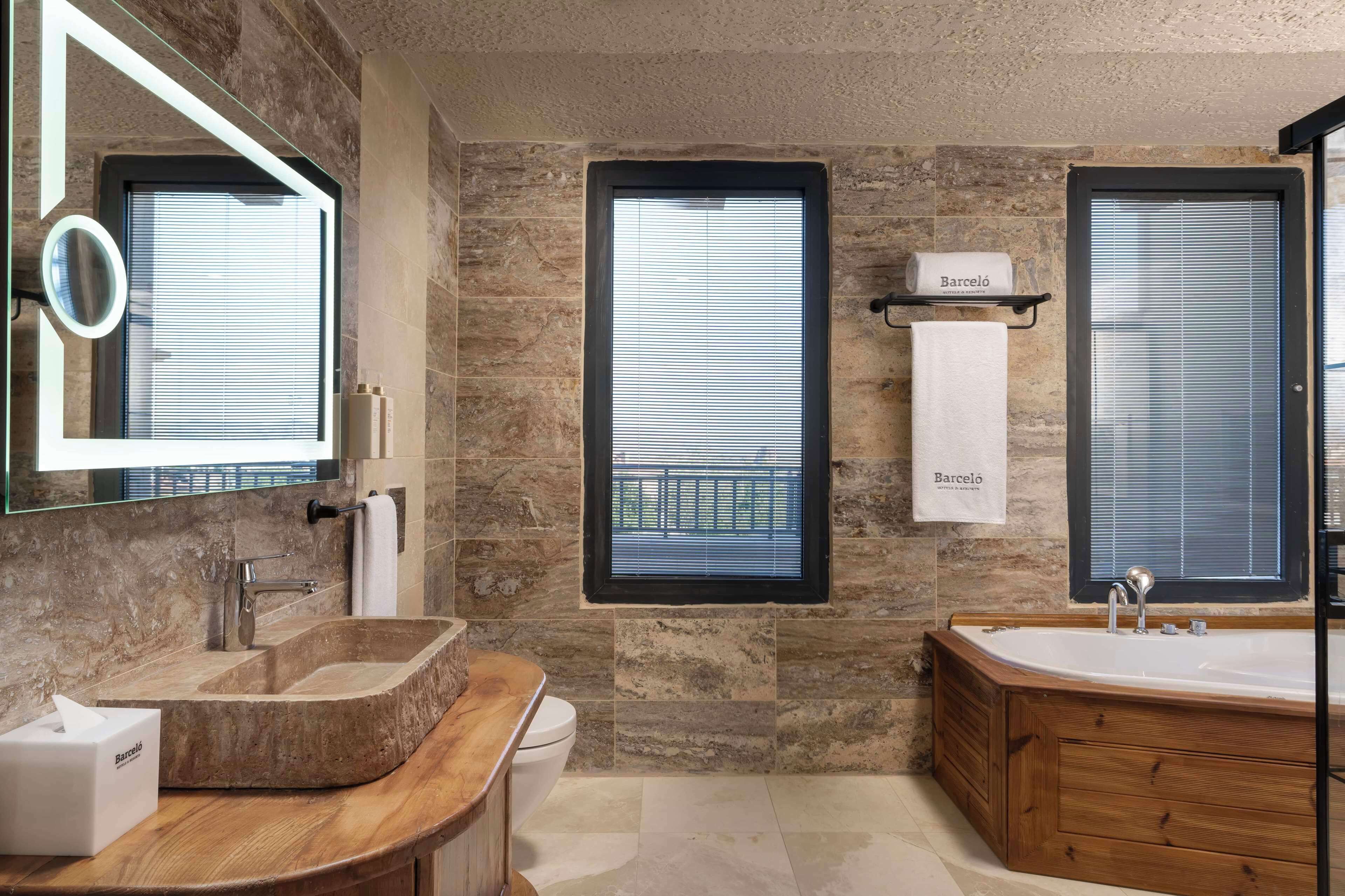 A modern bathroom with a stone wall, a freestanding bathtub, a wooden vanity, a large mirror, and two windows with blinds.