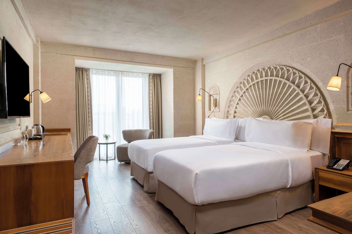 Ein Hotelzimmer mit zwei Betten, einem Schreibtisch, einem Sessel, einem kleinen Tisch und einem Fernseher.
