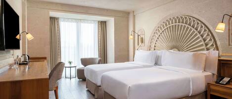 A hotel room with two beds, a desk, a chair, a small table, and a TV.